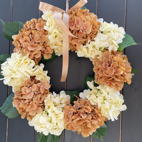 An artificial floral wreath made of large hydrangea blooms in peach, cream, and tan tones, with green leaves and a peach ribbon bow at the top, hanging against a dark gray wooden plank background.