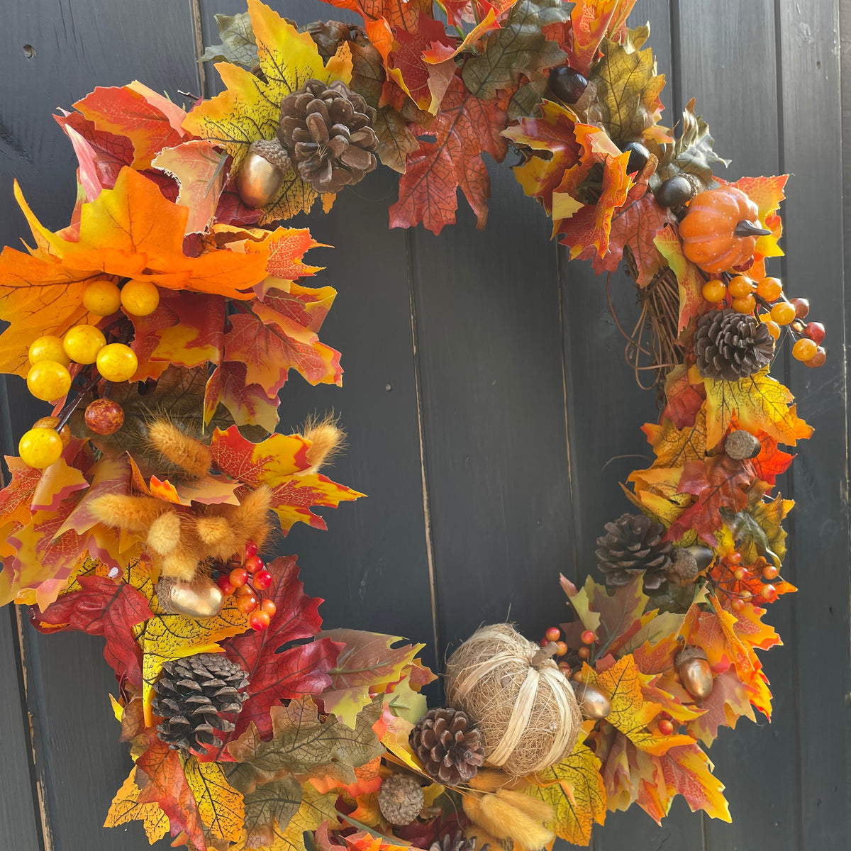 An autumn-themed decorative wreath with colorful artificial maple leaves in orange, yellow, red, and brown, decorated with pinecones, small pumpkins, acorns, and berry clusters, shown against a dark gray wooden plank background.