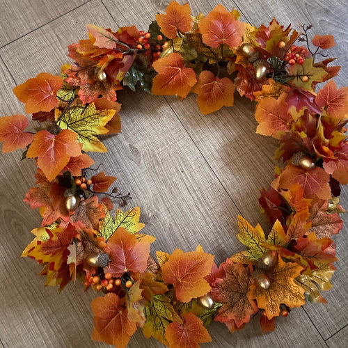 An autumn-themed artificial wreath made of orange, yellow, and red maple leaves, small berries, and gold-colored decorative beads, displayed on a light wood floor.