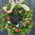 An artificial greenery wreath decorated with faux strawberries, white daisies, and a large beige burlap bow, displayed against a dark gray wooden plank background.