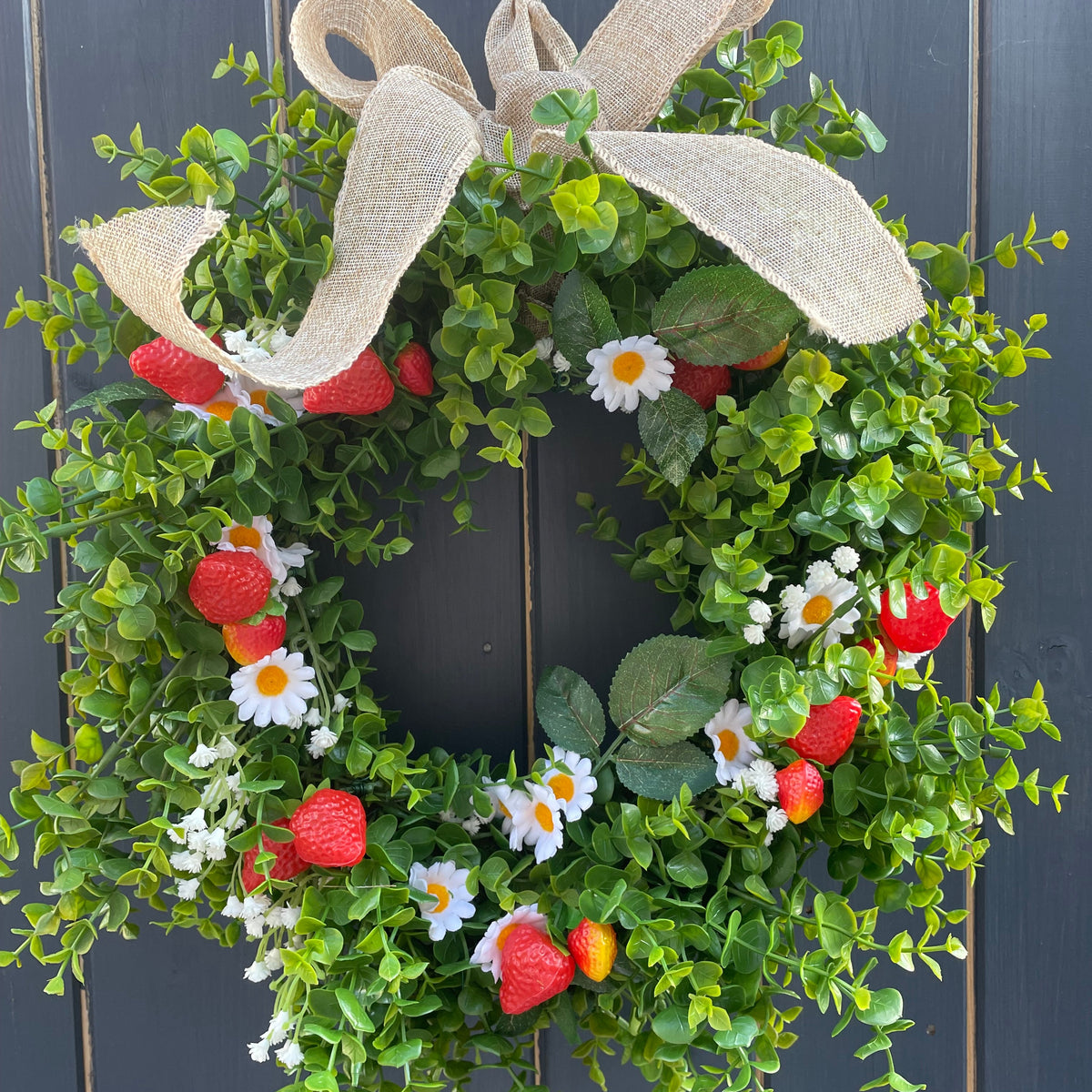 An artificial greenery wreath decorated with faux strawberries, white daisies, and a large beige burlap bow, displayed against a dark gray wooden plank background.