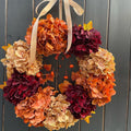 An autumn-themed floral wreath made of hydrangea blooms in shades of orange, peach, beige, and deep burgundy, accented with small orange berries and a cream ribbon bow, hanging against a dark gray wooden plank wall.
