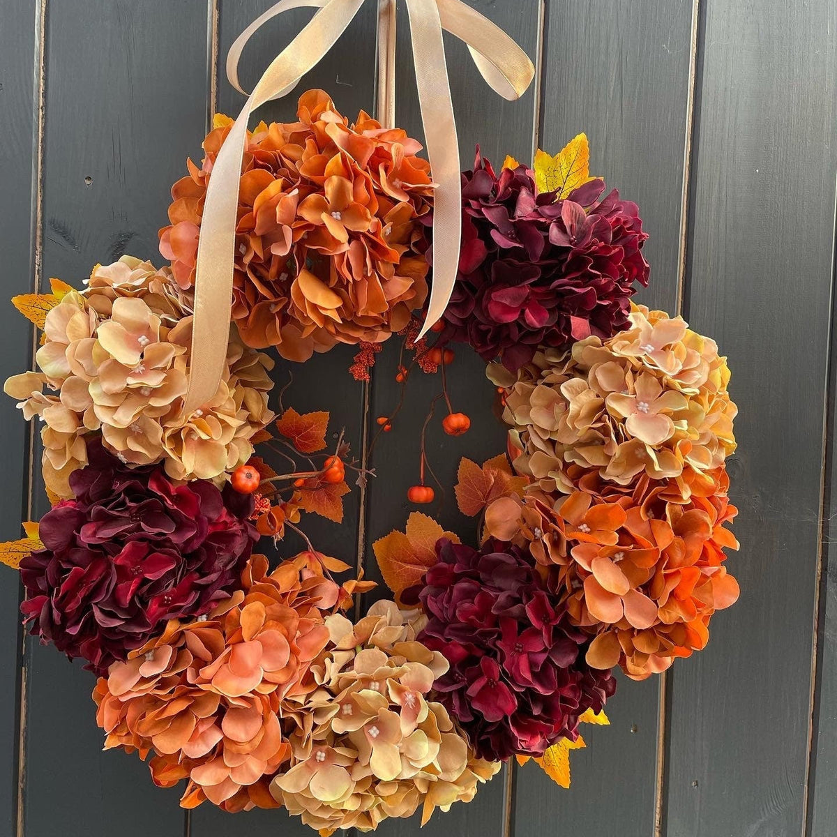 An autumn-themed floral wreath made of hydrangea blooms in shades of orange, peach, beige, and deep burgundy, accented with small orange berries and a cream ribbon bow, hanging against a dark gray wooden plank wall.
