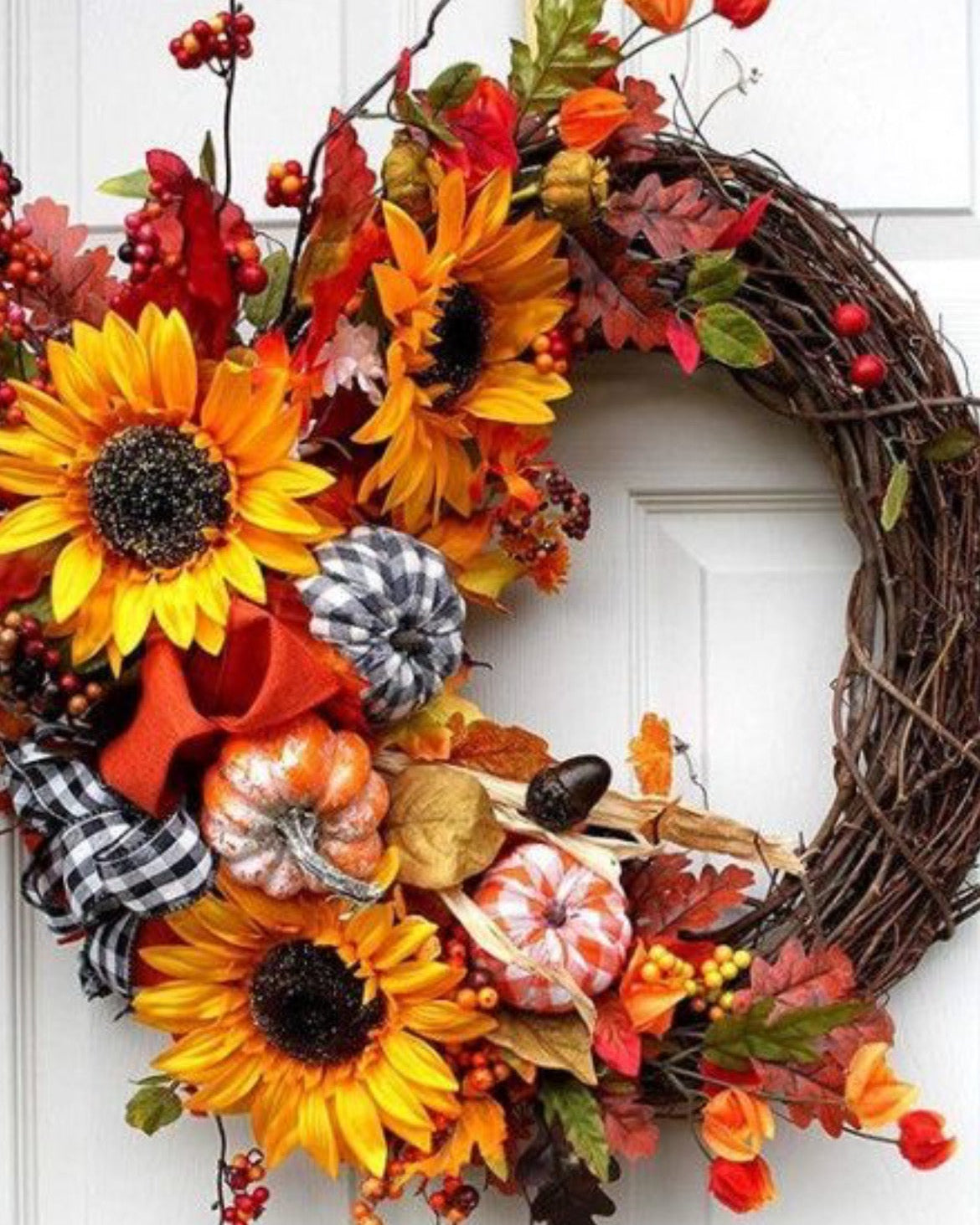 An autumn-themed grapevine wreath hanging on a white paneled door, decorated with large yellow sunflowers, orange and red fall leaves, small pumpkins, berries, and plaid ribbon.