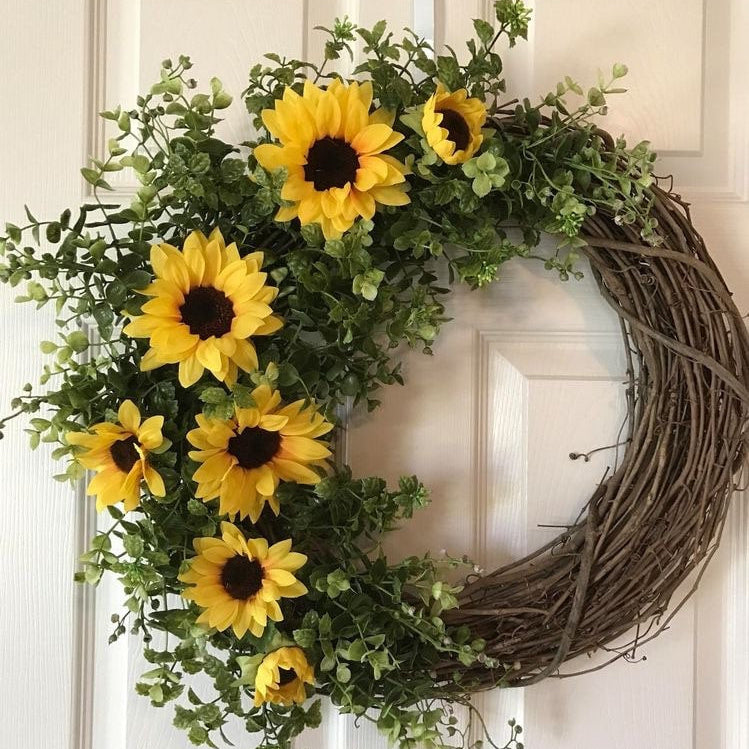 A rustic grapevine wreath decorated with bright yellow sunflowers and green foliage, hanging on a white paneled door.